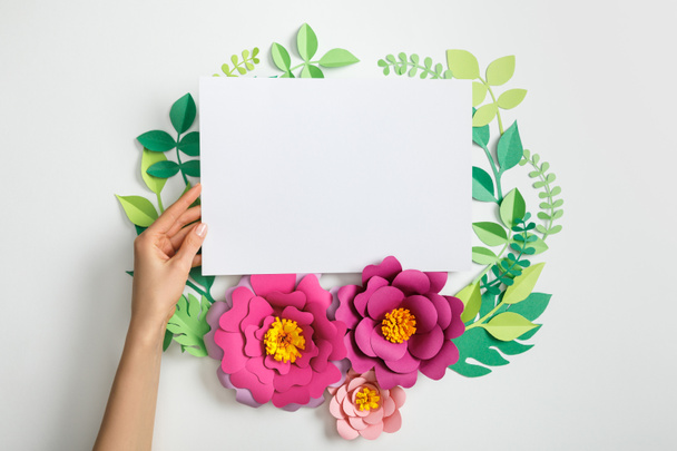 cropped view of woman putting white blank card near pink paper flowers and leaves on grey background - Photo, Image
