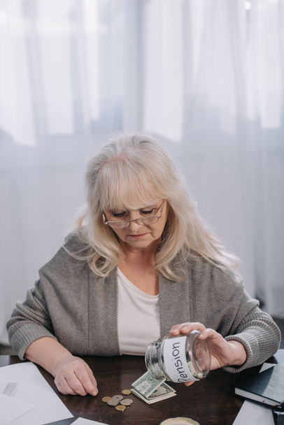 Senior femme tenant en pot de verre avec le mot « pension » en comptant l’argent à la maison - Photo, image