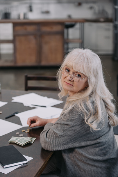 sad female pensioner sitting at table, looking at camera and counting money at home - Fotó, kép