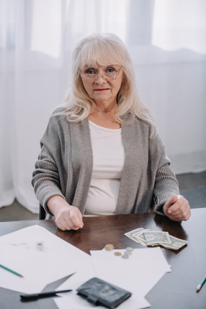 ανώτερος γυναίκα γυαλιά, κάθεται στο τραπέζι και κοιτάζοντας κάμερα ενώ μετρά τα χρήματα στο σπίτι - Φωτογραφία, εικόνα