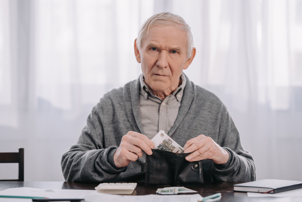 mężczyzna emeryt lub rencista siedząc przy stole i oddanie pieniędzy w portfelu, patrząc na kamery - Zdjęcie, obraz