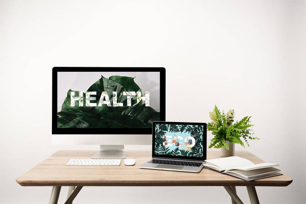 computer with health illustration and monstera leaves on monitor and laptop with eco lettering and fern leaves on screen on wooden table - Foto, Bild