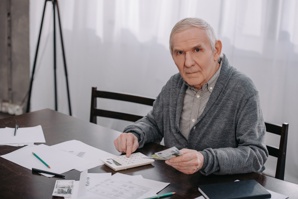 senior man in casual clothes sitting at table with paperwork and using calculator while counting money  - Foto, afbeelding