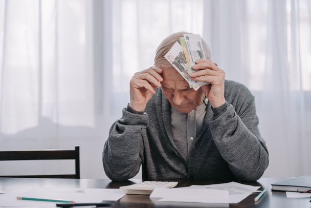 estresado hombre mayor sentado en la mesa con el papeleo y la celebración de dinero en casa
 - Foto, Imagen