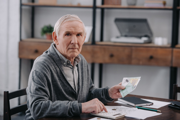hombre mayor sentado en la mesa, mirando a la cámara, usando la calculadora y contando dinero
 - Foto, Imagen