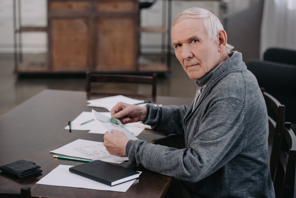 starší muž, sedící u stolu s papíry, při pohledu na fotoaparát a drží obálku s penězi  - Fotografie, Obrázek