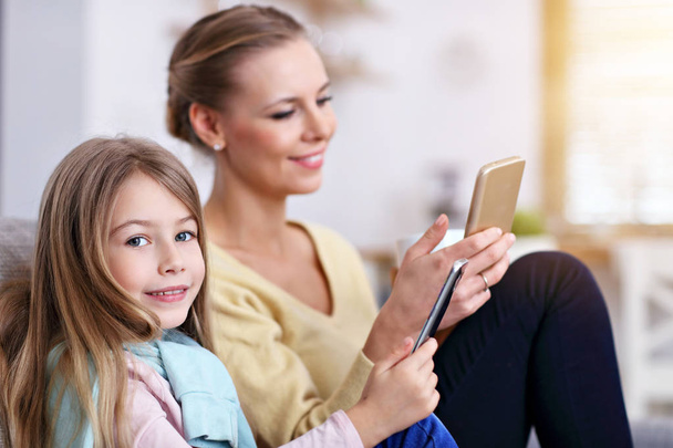 Νεαρή μαμά και η μικρή κόρη της χρησιμοποιώντας το smartphone και κάθεται στον καναπέ στο σπίτι - Φωτογραφία, εικόνα