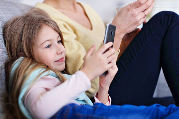Joven mamá y su hija pequeña utilizando el teléfono inteligente y sentado en el sofá en casa
 - Foto, imagen