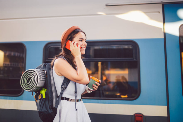 theme tourism and travel young student. beautiful young Caucasian girl in dress and hat standing at train station near train with backpack and cup coffee in hand. Uses smartphone phone technology - Photo, Image
