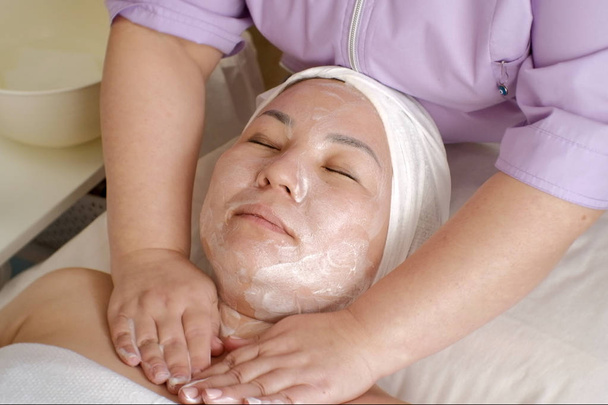 As mãos de um cosmetologist profissional põem o sabão suds no pescoço fêmea do paciente. Uma mulher de aparência asiática com olhos fechados em um procedimento de limpeza de pele de cara em um salão de beleza. Close-up
. - Foto, Imagem
