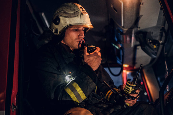 The fire brigade arrived at the night-time. Fireman sitting in the fire truck and talking on the radio - Photo, Image
