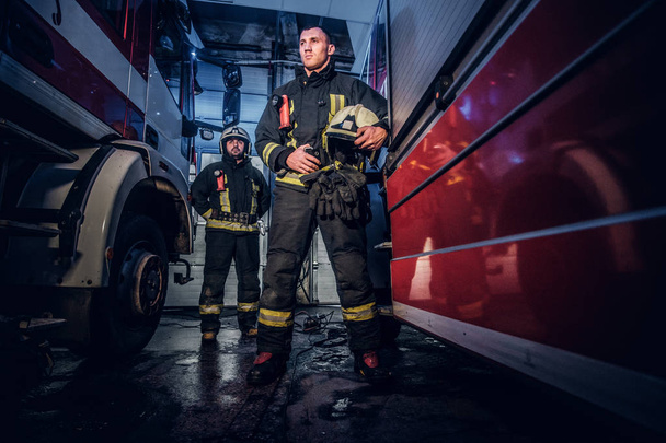 Ολόσωμο πορτρέτο από δύο γενναίους πυροσβέστες στην προστατευτική στολή στέκεται μεταξύ δύο πυροσβεστικά οχήματα στο γκαράζ του ο σταθμός πυρκαγιάς - Φωτογραφία, εικόνα