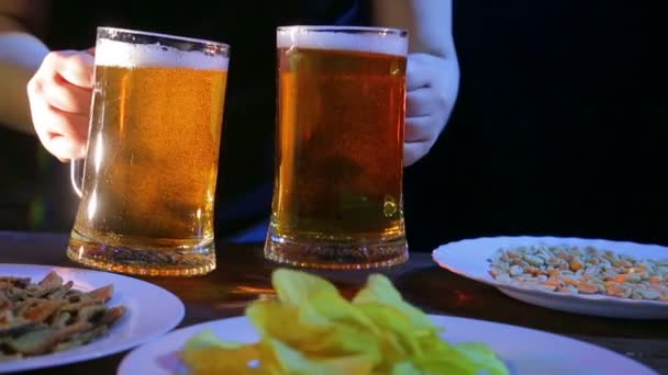 A female bartender puts on a table with snacks on a black background two full mugs of foaming beer - Séquence, vidéo
