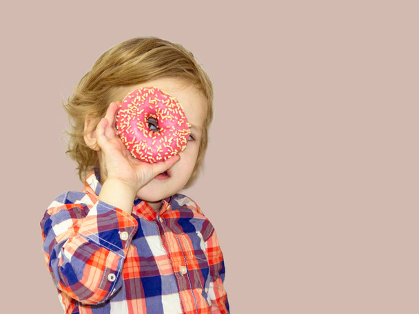 Little happy cute boy is eating donut on brown background wall. Child is having fun with donut. Tasty food for playing kids. Funny time  with sweet food. Bright baby boy in a plaid shirt. - Foto, Bild