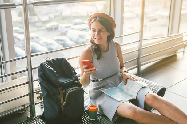 a turizmus és a fiatal diák utazási téma. Gyönyörű fiatal kaukázusi lány ruhában és kalapban ül a padlón turisztikai szőnyeg belsejében a repülőtéri terminál. Váróterem késleltetett repülés, késleltetés indulás - Fotó, kép