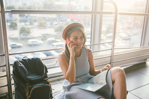 theme of tourism and travel of young student. Beautiful young caucasian girl in dress and hat sits on floor tourist rug inside terminal airport terminal. Waiting room delayed flight, delay departure - Foto, Imagen