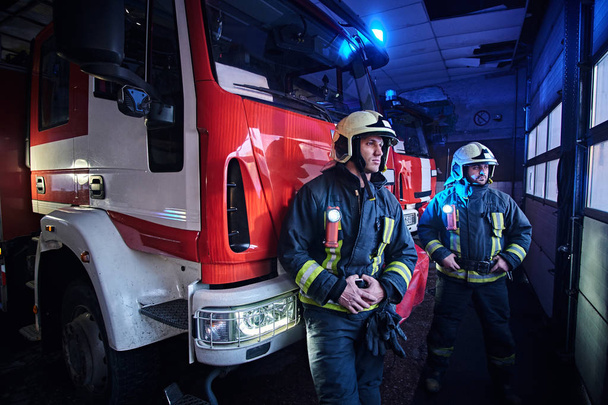 Δύο πυροσβέστες φορούσε προστατευτική στολή στέκεται δίπλα σε ένα πυροσβεστικό όχημα σε ένα γκαράζ του μια πυροσβεστική. - Φωτογραφία, εικόνα