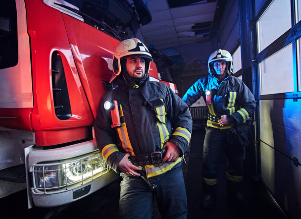 zwei Feuerwehrmänner in Schutzuniform stehen neben einem Feuerwehrauto in einer Garage einer Feuerwehr. - Foto, Bild