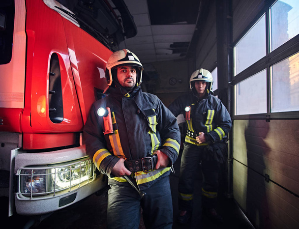 Dois bombeiros vestindo uniforme de proteção ao lado de um caminhão de bombeiros em uma garagem de um corpo de bombeiros
. - Foto, Imagem