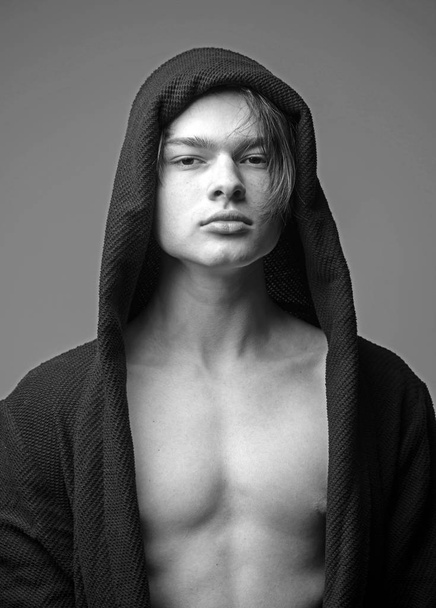 Guy on confident face in black hood, grey background. Masculinity concept. Macho in black bathrobe with hood, close up. Man with smooth skin on face, neck and chest looking at camera. - Zdjęcie, obraz