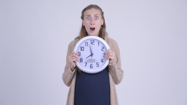 Young stressed pregnant woman holding wall clock and looking shocked - Imágenes, Vídeo