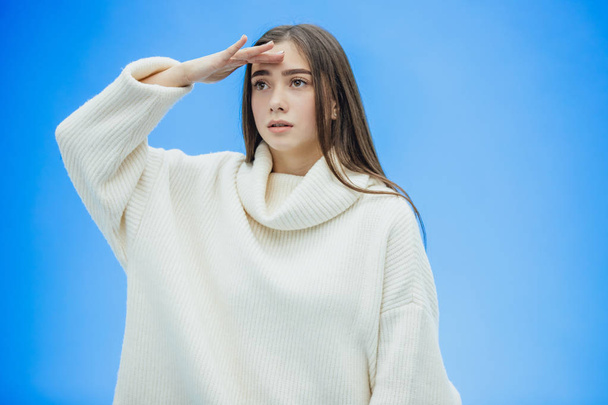 A young woman wearing a winter sweater on an isolated blue background, without smiling, raising her hand up from pain puts them on the forehead. - Photo, Image