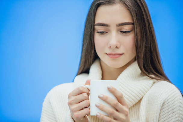 Interestingly, a young woman in a knitted white sweater. Holds a white cup of coffee or tea in his hands. Isolated against the background of a blue wall, studio portrait. Concept of a way of life of - Fotoğraf, Görsel