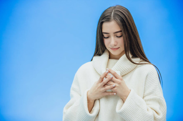 Interestingly, a young woman in a knitted white sweater. Holds a white cup of coffee or tea in his hands. Isolated against the background of a blue wall, studio portrait. Concept of a way of life of - Foto, Bild