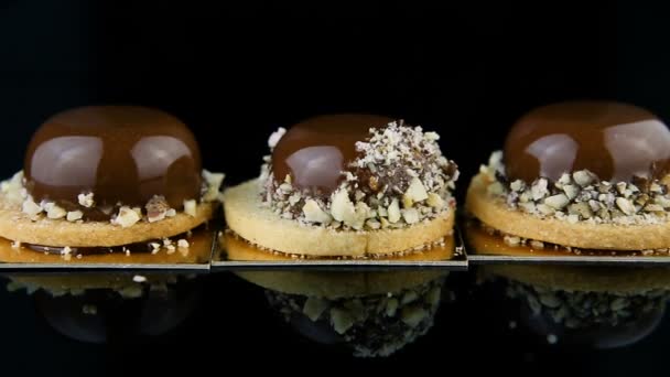 ομαλό ζουμ έξω από τρία Γαλλικά μίνι μους γλυκά ζύμης καλυμμένο με γλάσο σοκολάτας και διακοσμημένα με θρυμματισμένα καρύδια που σερβίρεται σε φόντο μαύρο καθρέφτη - Πλάνα, βίντεο