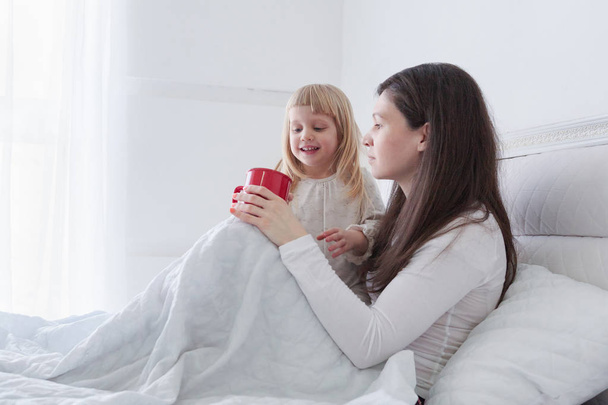 Young mother with daughter drinking some tea while sitting in bed in white bedroom - Foto, Imagen
