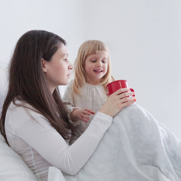 Young mother with daughter drinking some tea while sitting in bed in bedroom - Zdjęcie, obraz