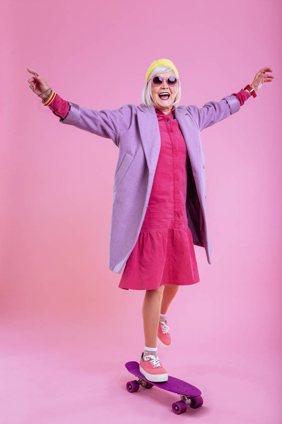 Retired woman wearing pink dress standing on purple skateboard - Zdjęcie, obraz