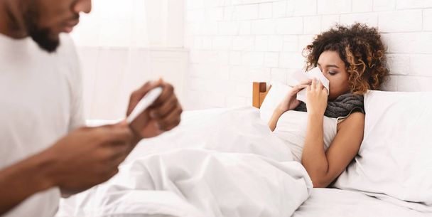 Sick woman in bed, man checking temperature on thermometer - Foto, afbeelding