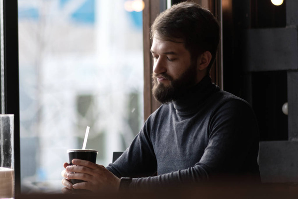 attractive young stylish man sitting in a cafe and looking out the window. Business, freelance. pensive bearded guy  near the window in a bar - Foto, imagen
