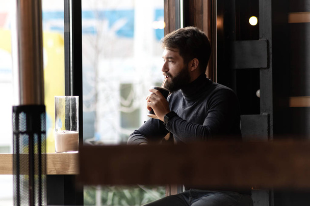 attraktiver junger stilvoller Mann sitzt in einem Café und schaut aus dem Fenster. Geschäft, freiberuflich. Nachdenklicher bärtiger Typ am Fenster einer Bar - Foto, Bild