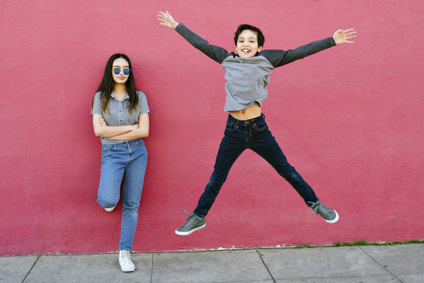 Um garotinho tenta impressionar sua irmã mais velha pulando alto. Ela está contra uma parede urbana sem impressionar
 - Foto, Imagem