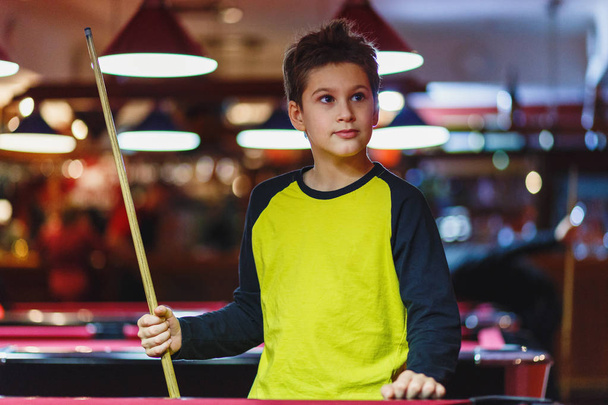 Cute boy in yellow t shirt plays billiard or pool in club. Young Kid learns to play snooker. Boy with billiard cue strikes the ball on table. Active Leisure, sport, hobby concept - Фото, изображение