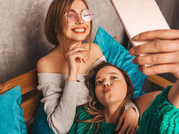 Two young beautiful smiling gorgeous girls in trendy summer hipster clothes.  Sexy carefree women posing in interior and taking selfie. Positive models having fun with smartphone - Fotoğraf, Görsel