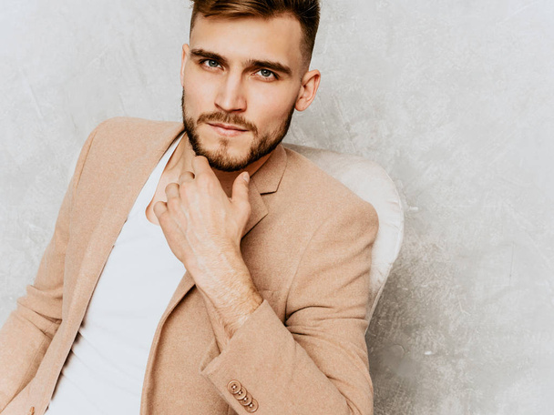 Portrait of handsome smiling hipster lumbersexual businessman model wearing casual beige suit. Fashion stylish man posing against gray wall sitting in chair in interior - Φωτογραφία, εικόνα