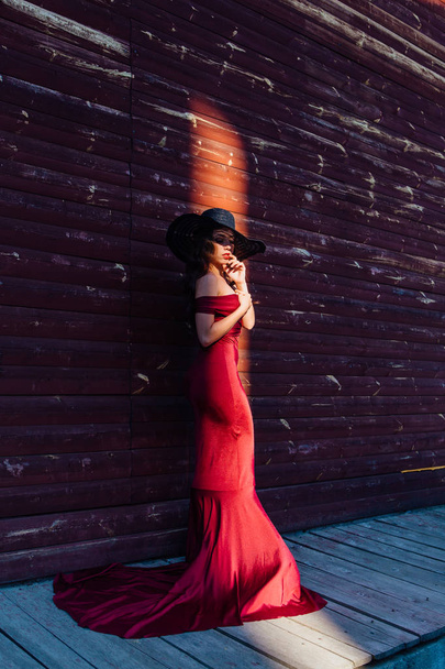Beautiful young woman in a long red evening gown with a train and black hat standing in ray of light - Фото, зображення