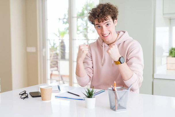 Young student man writing on notebook and studying very happy and excited doing winner gesture with arms raised, smiling and screaming for success. Celebration concept. - Фото, изображение
