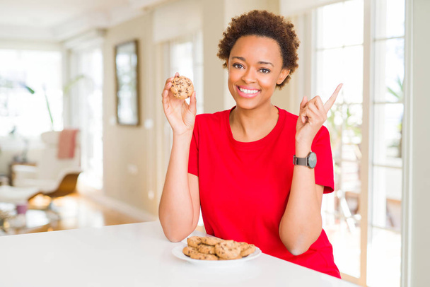 Joven afroamericana americana comiendo galletas fritas de chocolate muy feliz señalando con la mano y el dedo a un lado
 - Foto, Imagen