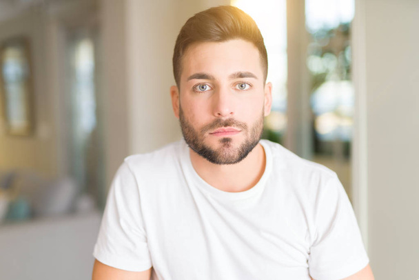 Young handsome man wearing casual white t-shirt at home with serious expression on face. Simple and natural looking at the camera. - Photo, Image