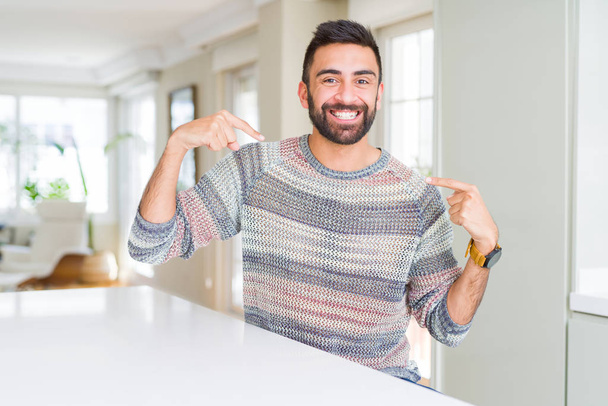 bel homme hispanique portant pull décontracté à la maison regardant confiant avec le sourire sur le visage, se montrant du doigt fier et heureux
. - Photo, image