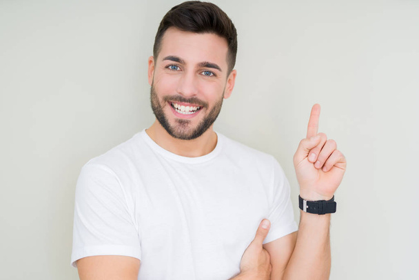 Jonge knappe man dragen casual wit t-shirt over geïsoleerde achtergrond met een grote glimlach op het gezicht, met de hand en de vinger te wijzen naar de kant te kijken naar de camera. - Foto, afbeelding