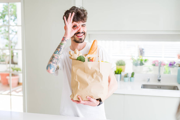 Молодий чоловік тримає паперовий мішок свіжих продуктів з супермаркету з щасливим обличчям, посміхаючись, роблячи знак ОК з рукою на око, дивлячись через пальці
 - Фото, зображення