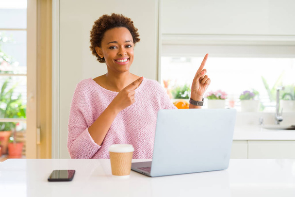 Молодая африканская американка, работающая с помощью компьютера, улыбается и смотрит в камеру, указывая двумя руками и пальцами в сторону.
. - Фото, изображение