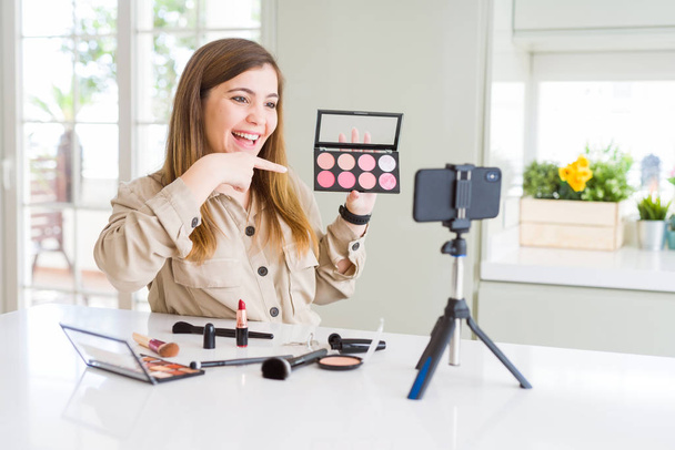 Mooie jonge beïnvloeder vrouw opname make-up video tutorial erg blij wijzen met hand en vinger - Foto, afbeelding
