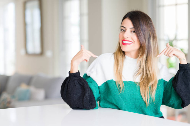 Mujer hermosa joven vistiendo suéter de invierno en casa mirando confiado con sonrisa en la cara, señalándose con los dedos orgullosos y felices
. - Foto, imagen