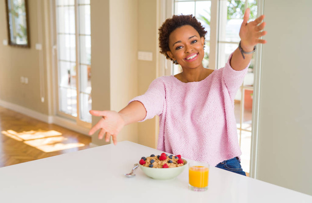 Nuori afrikkalainen amerikkalainen nainen nauttii terveellistä aamiaista kotona katsoen kameraa hymyillen avosylin halaten. Iloinen ilme, johon kuuluu onnellisuus
. - Valokuva, kuva
